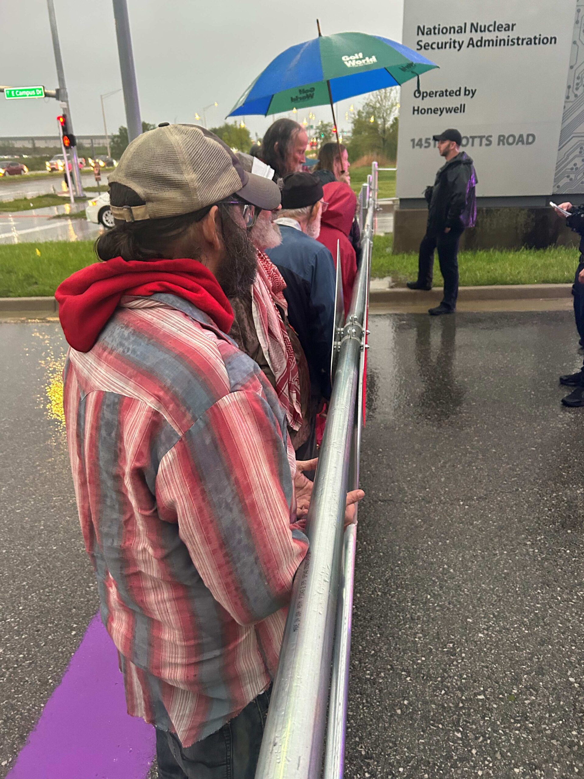 Activists line up along a guardrail in the rain beside a sign reading "National Nuclear Security Administration / Operated by Honeywell"; a bearded man in a red plaid shirt is in front.