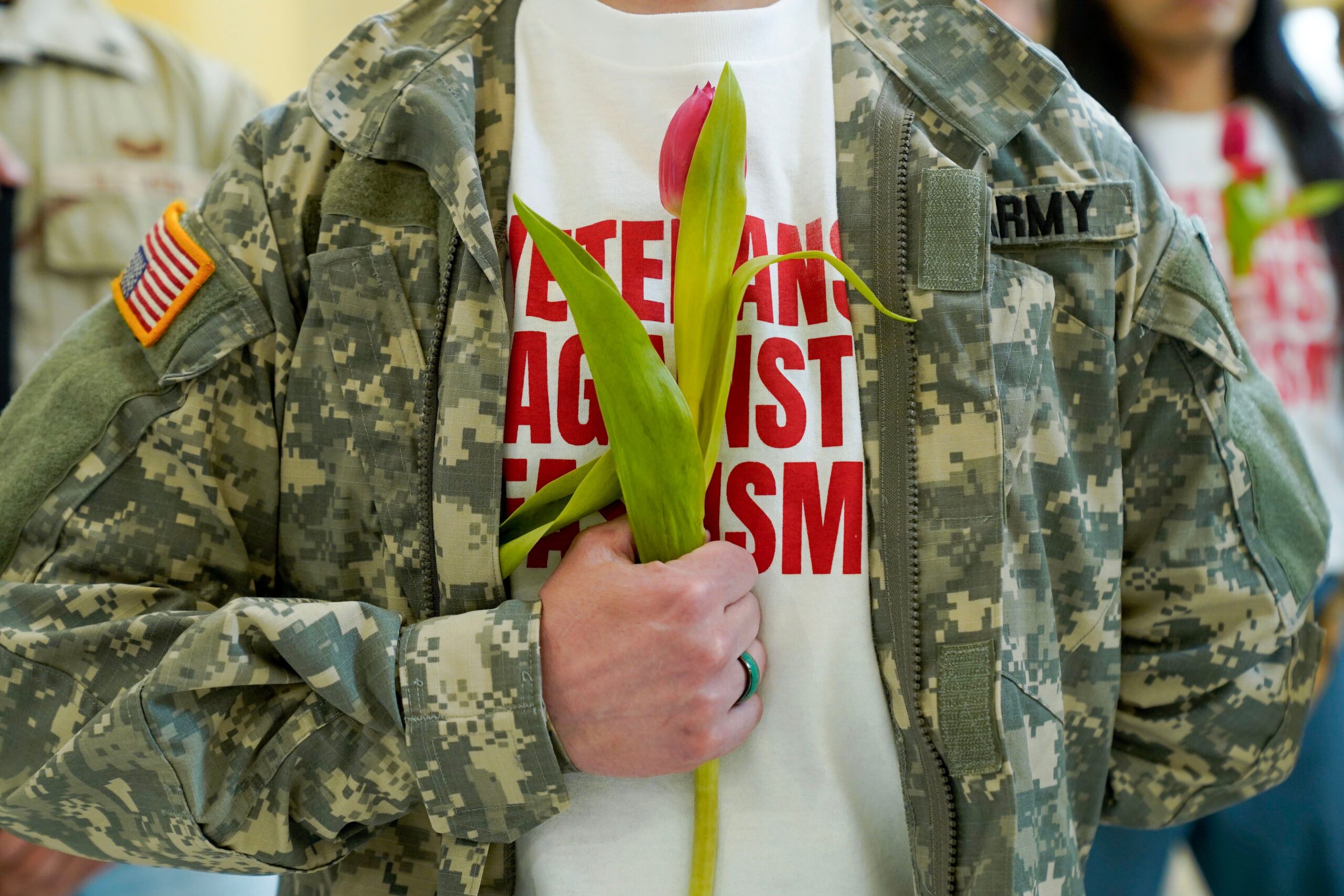 Close-up of a person in a U.S. Army camouflage jacket and a "VETERANS AGAINST FASCISM" T-shirt holding a single yellow tulip against their chest.
