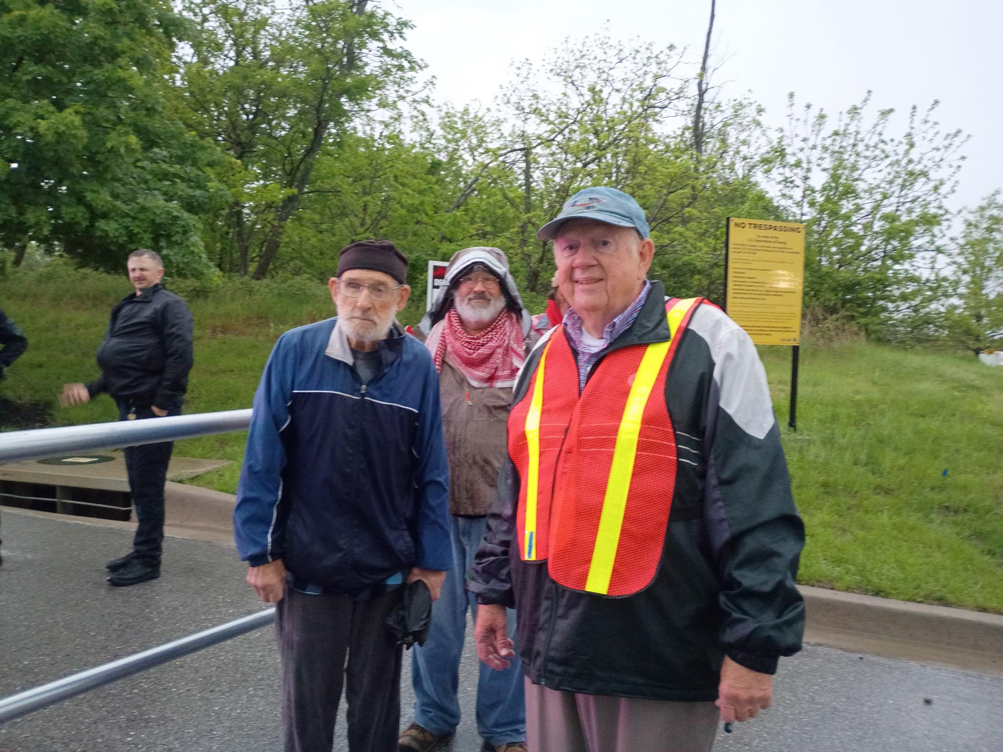 Three older activists pose together in the rain at a roadside; the man in front wears an orange safety vest, a yellow plant sign is behind them.