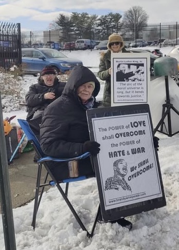 Bundled-up activists sit in folding chairs on a snowy roadside; one holds a placard reading "The power of LOVE shall overcome the power of HATE & WAR."