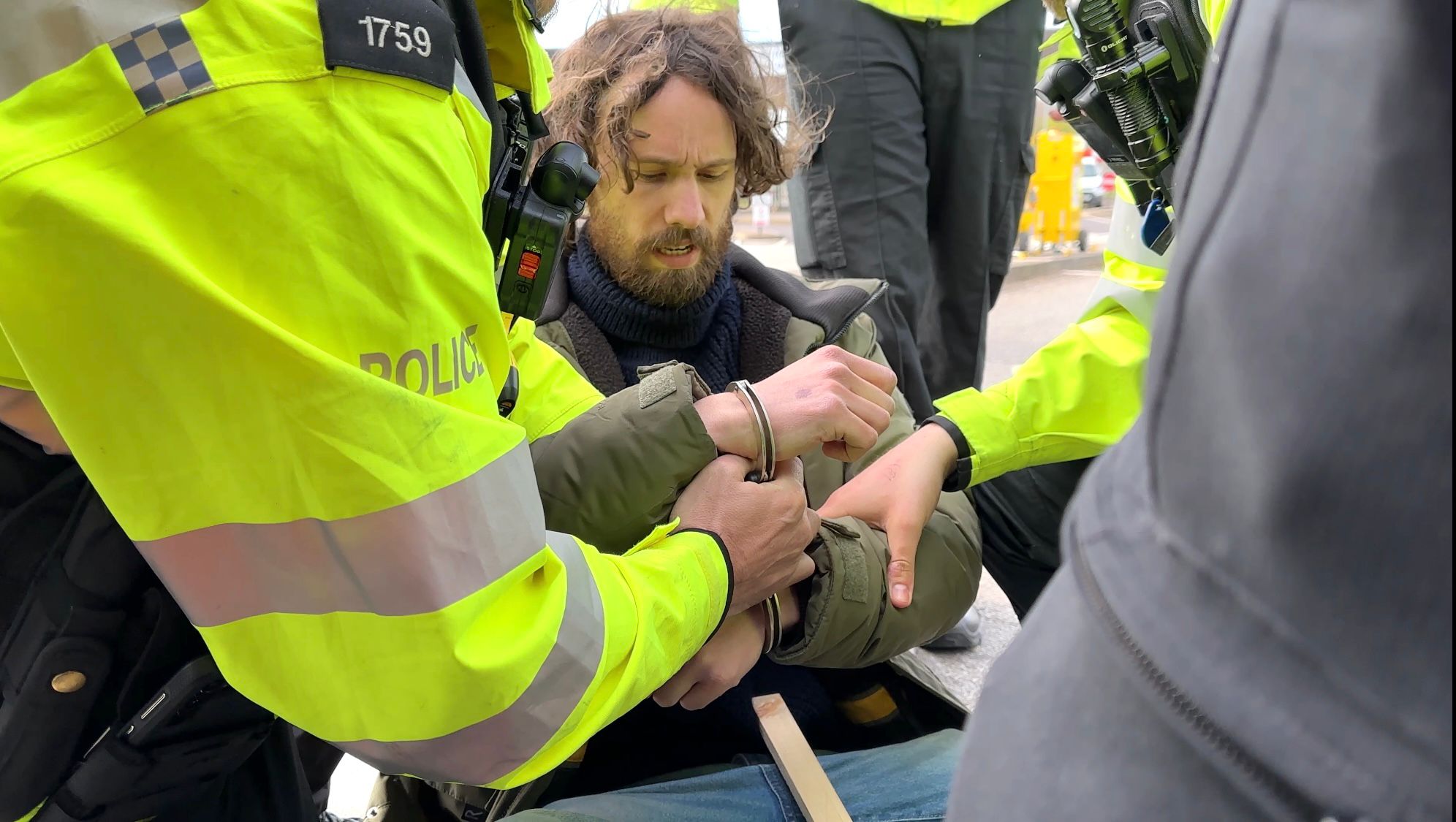 Yellow-jacketed police handcuff a bearded man in a green jacket who is seated on the ground during a roadside arrest.