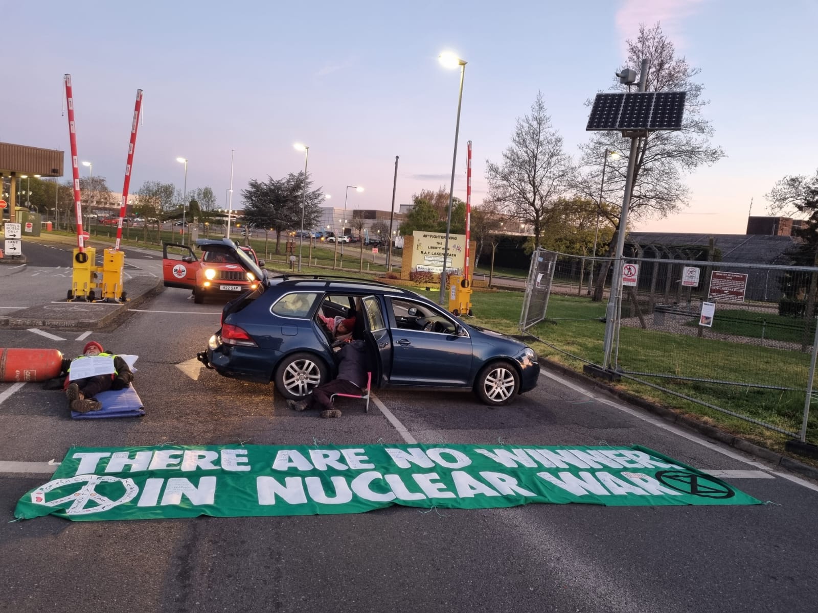 At dusk, a green car is parked blocking a base gate with a green banner laid on the road reading "THERE ARE NO WINNERS IN NUCLEAR WAR."