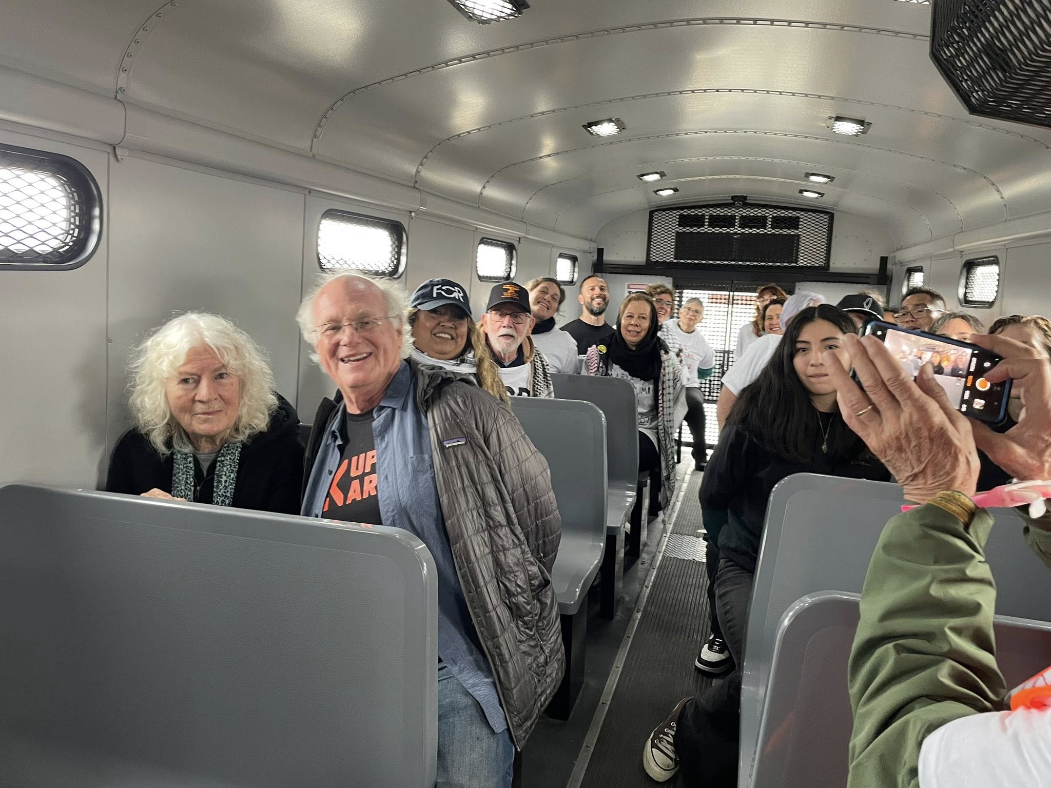 Older activists sit on a curb being processed by police; one smiles and waves while another raises a phone to take a photo.