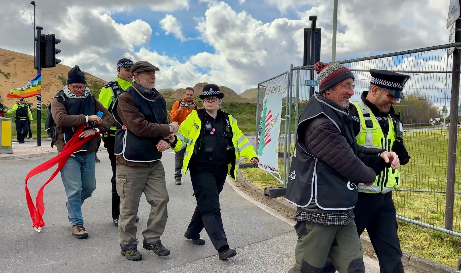 Activists trailing red ribbon walk along a fence line as police officers escort and follow them.