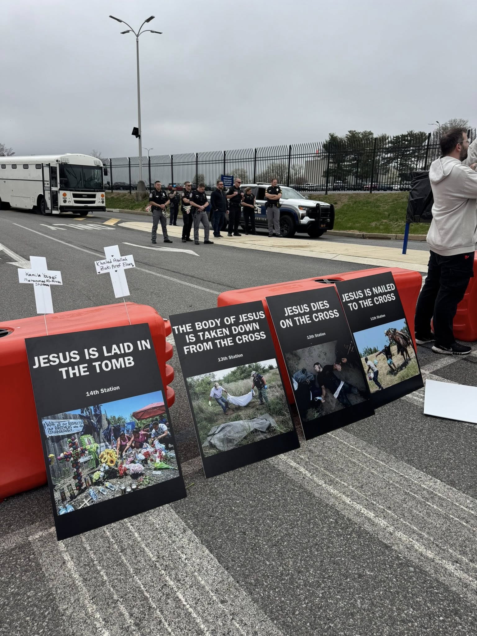 A row of placards on the ground depicting Stations of the Cross, including "JESUS IS LAID IN THE TOMB," with small white crosses, as police and a vehicle gather behind.