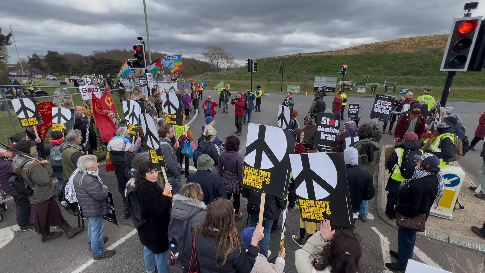 A crowd of protesters fills a road at a base entrance holding many black-and-white peace-symbol placards under a cloudy sky.