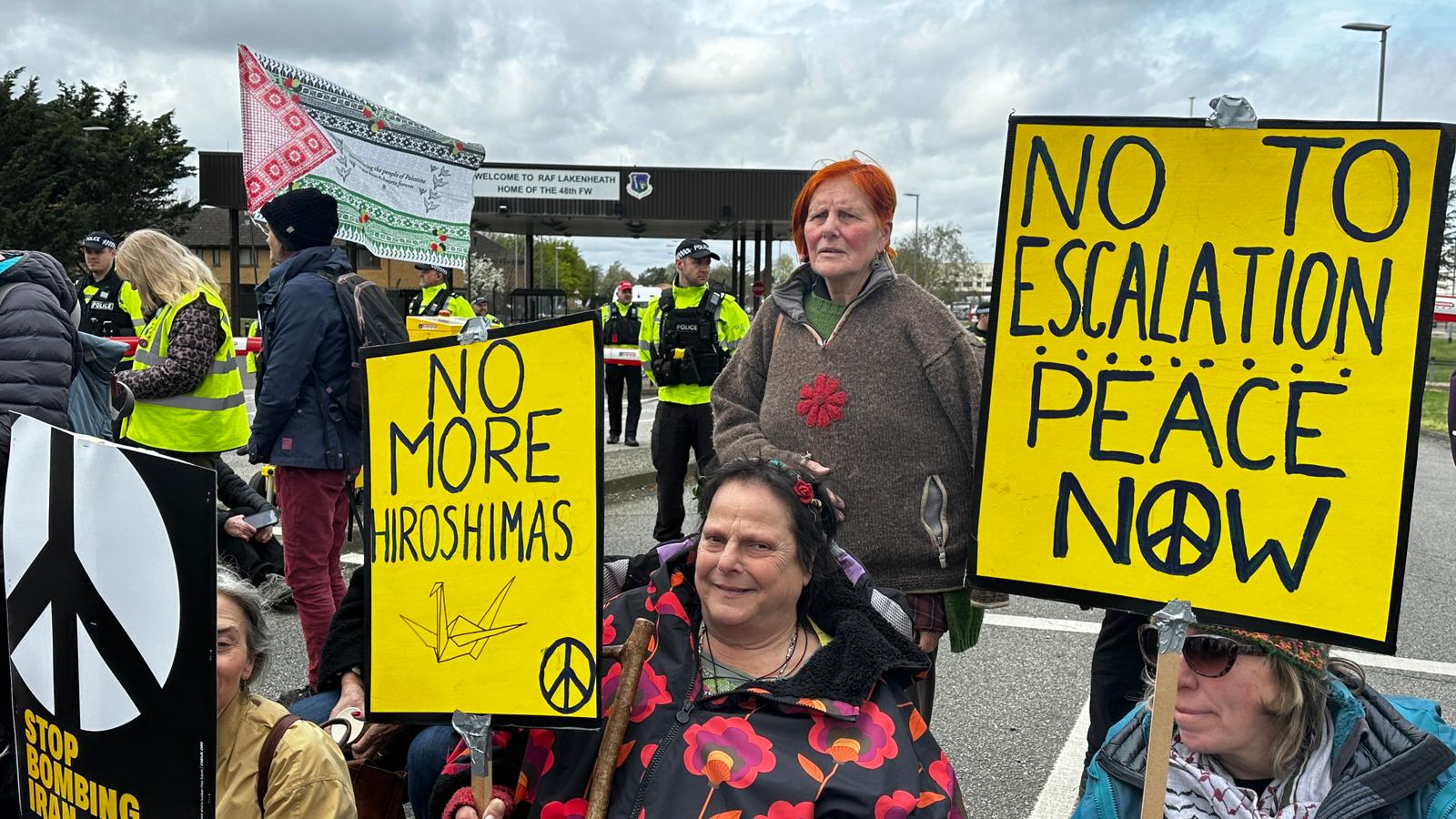 Protesters at a base gate hold large yellow signs reading "NO MORE HIROSHIMAS" and "NO TO ESCALATION — PEACE NOW," some seated in wheelchairs.