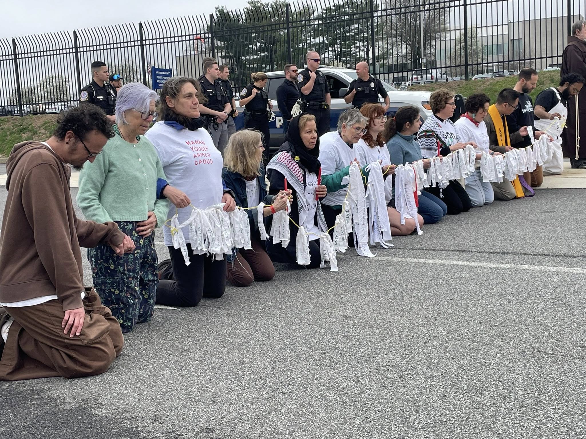 A row of kneeling activists holds a long line of white cloth banners across a roadway while police officers stand behind them.