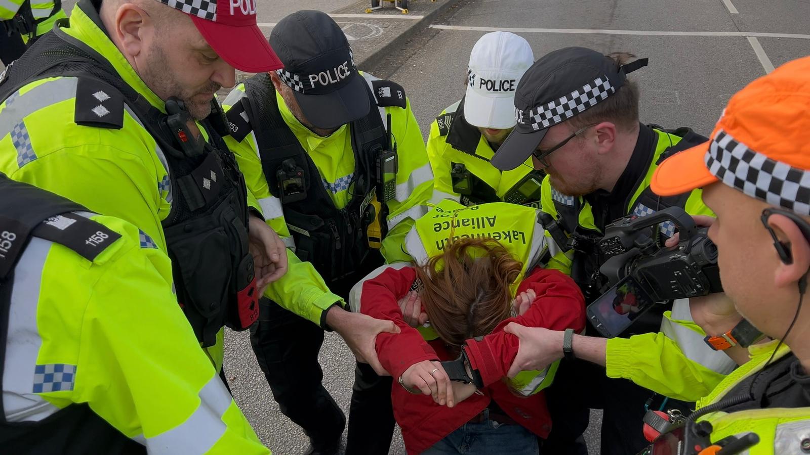 Several yellow-jacketed police officers lift and restrain a person in a red jacket on the road during an arrest.