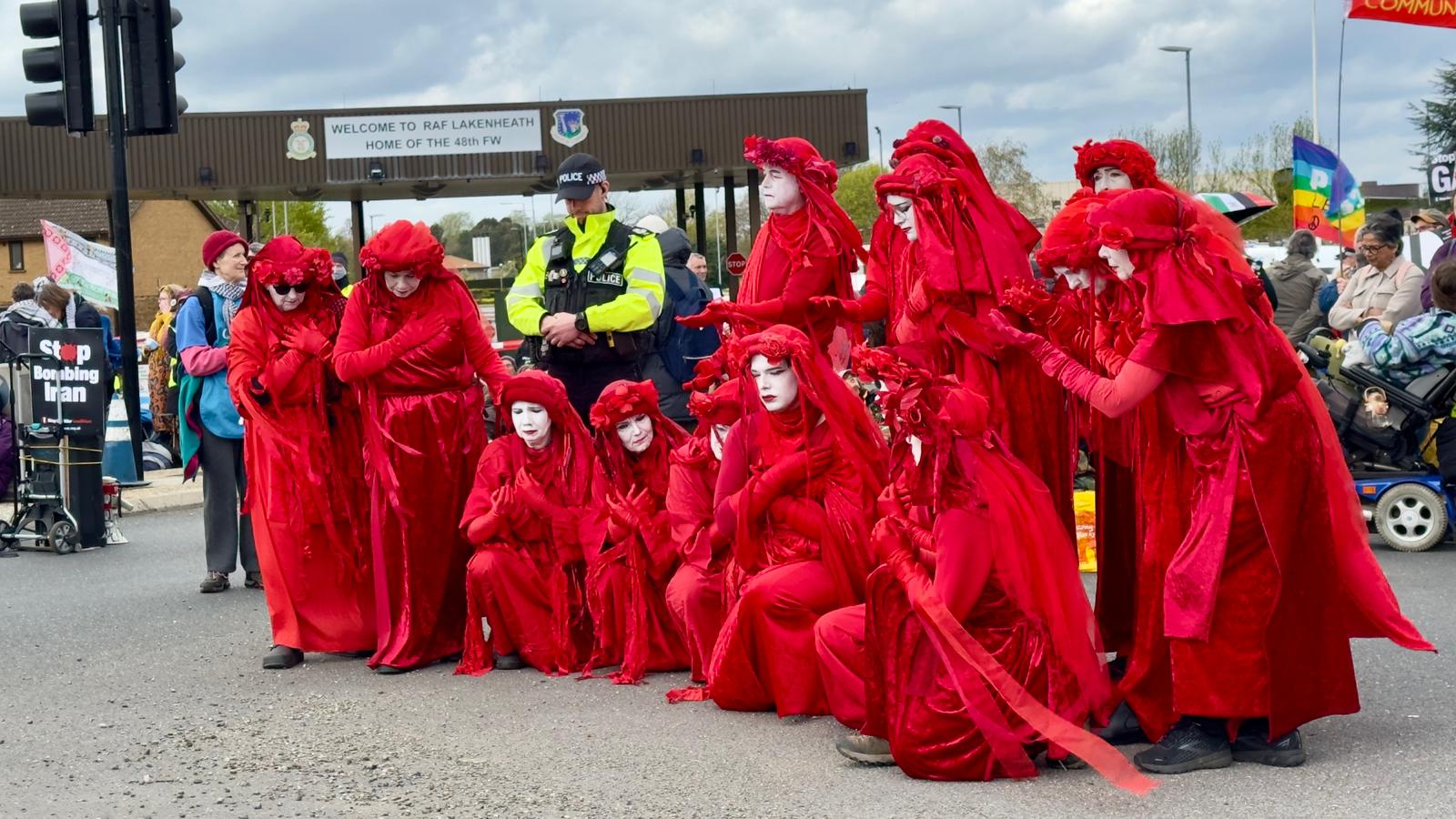 A group of activists dressed in flowing red robes and white face paint (Extinction Rebellion "Red Rebel Brigade" style) stand in a line at a protest as a police officer looks on.