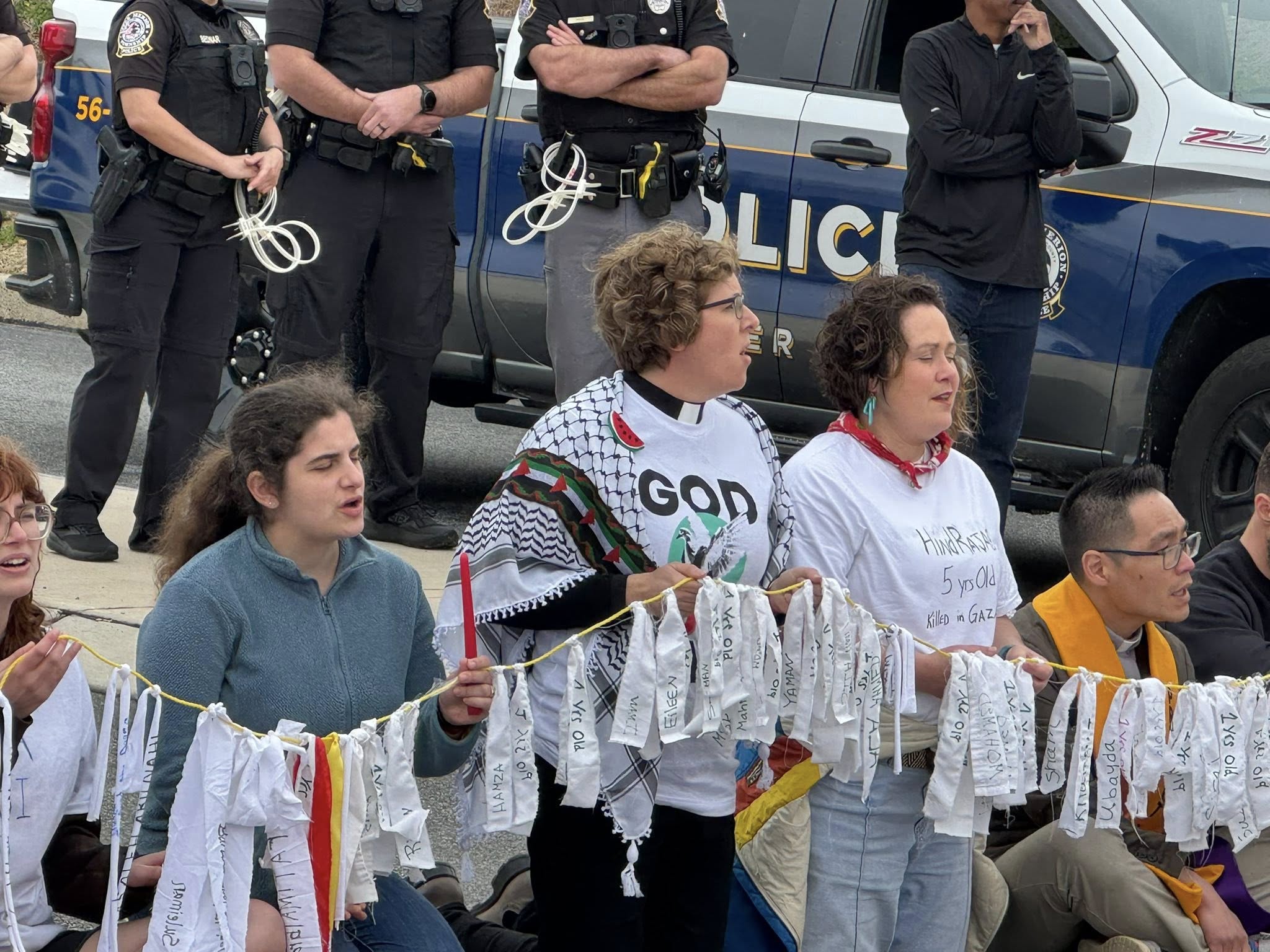Activists sit on the ground holding a long string of small white cloth banners; one wears a keffiyeh and a "GOD" sign, with police standing behind.
