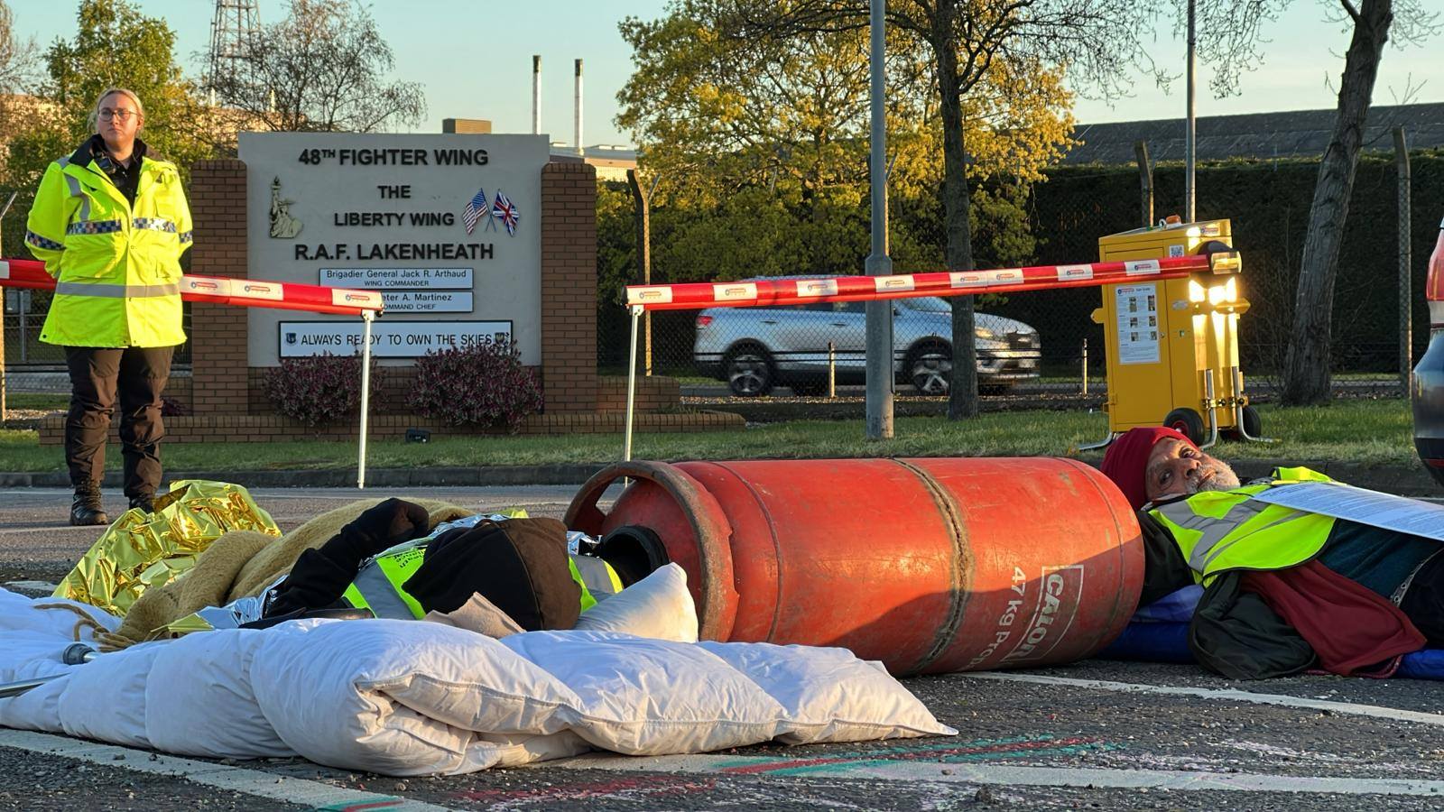 Activists lie across a base access road locked to a large orange tube blockade device; a vested steward stands by a barrier sign reading "THE LIBERTY MOB / RAF LAKENHEATH."