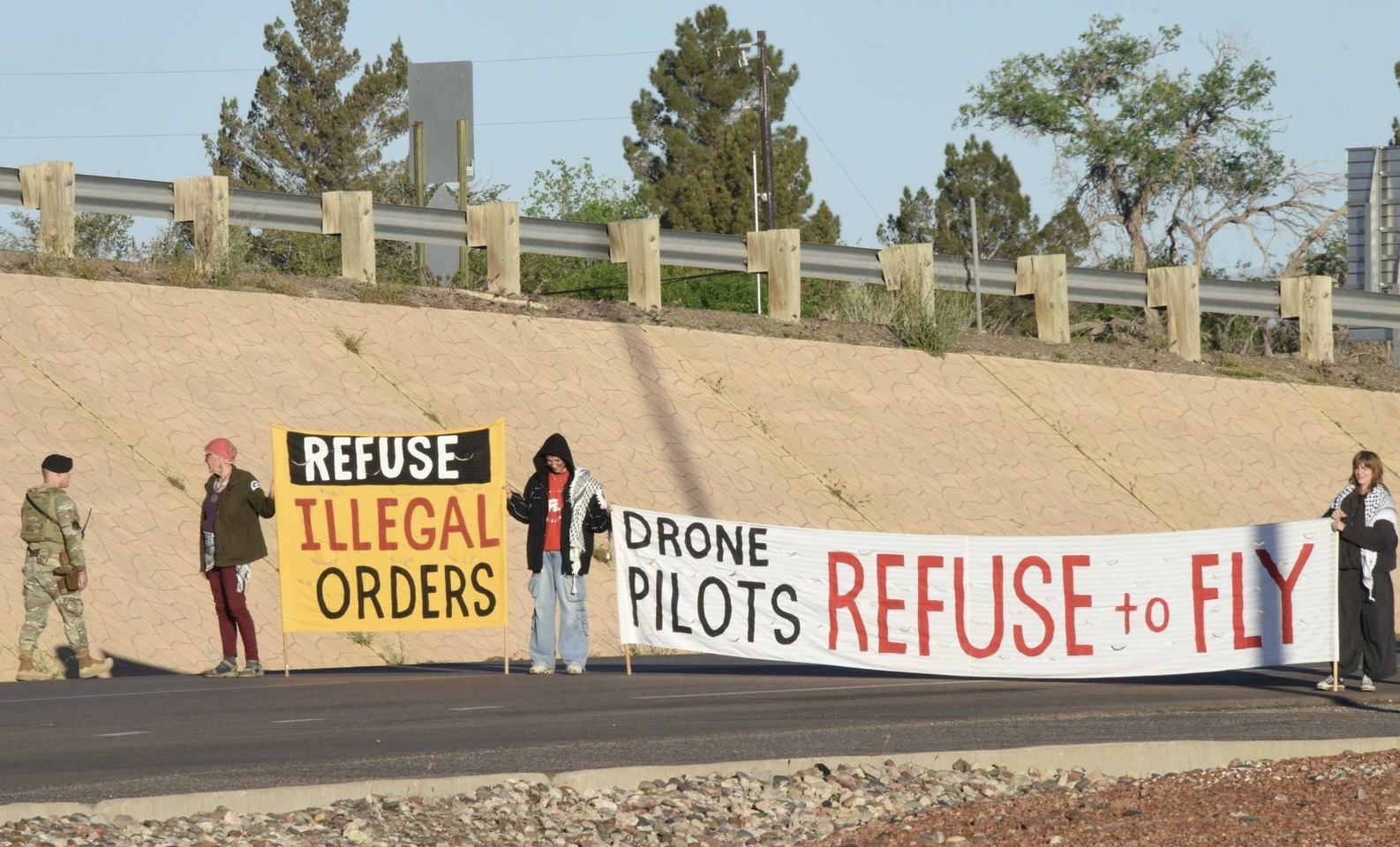 Activists hold a banner reading "REFUSE ILLEGAL ORDERS — DRONE PILOTS REFUSE TO FLY" along a desert roadside as a uniformed servicemember stands at left.