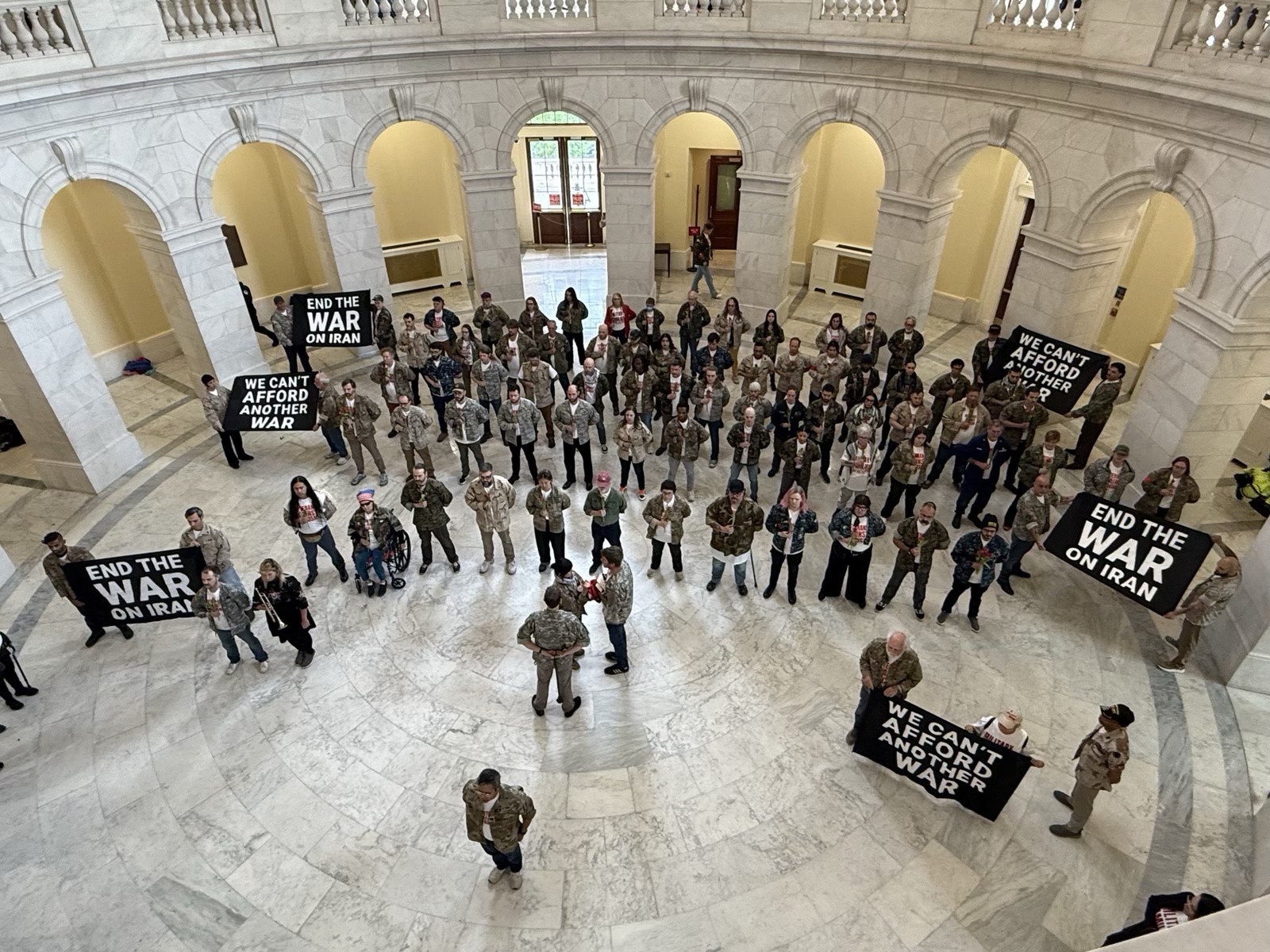 Overhead view of dozens of veterans in fatigues standing in a marble rotunda holding black banners reading "END THE WAR ON IRAN" and "WE CAN'T AFFORD ANOTHER WAR."