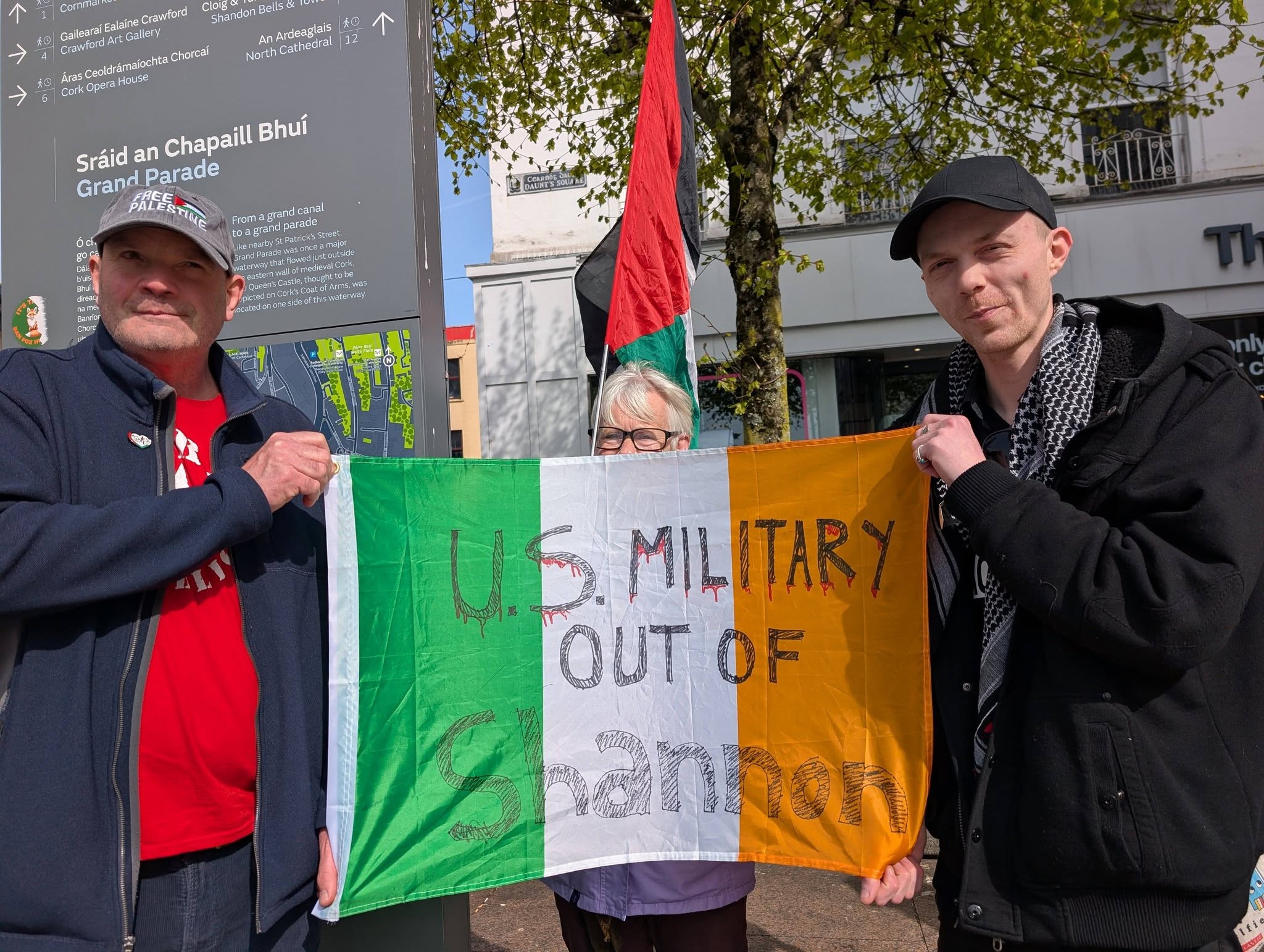 Three people hold an Irish tricolour flag hand-painted "U.S. MILITARY OUT OF SHANNON" in a town square.