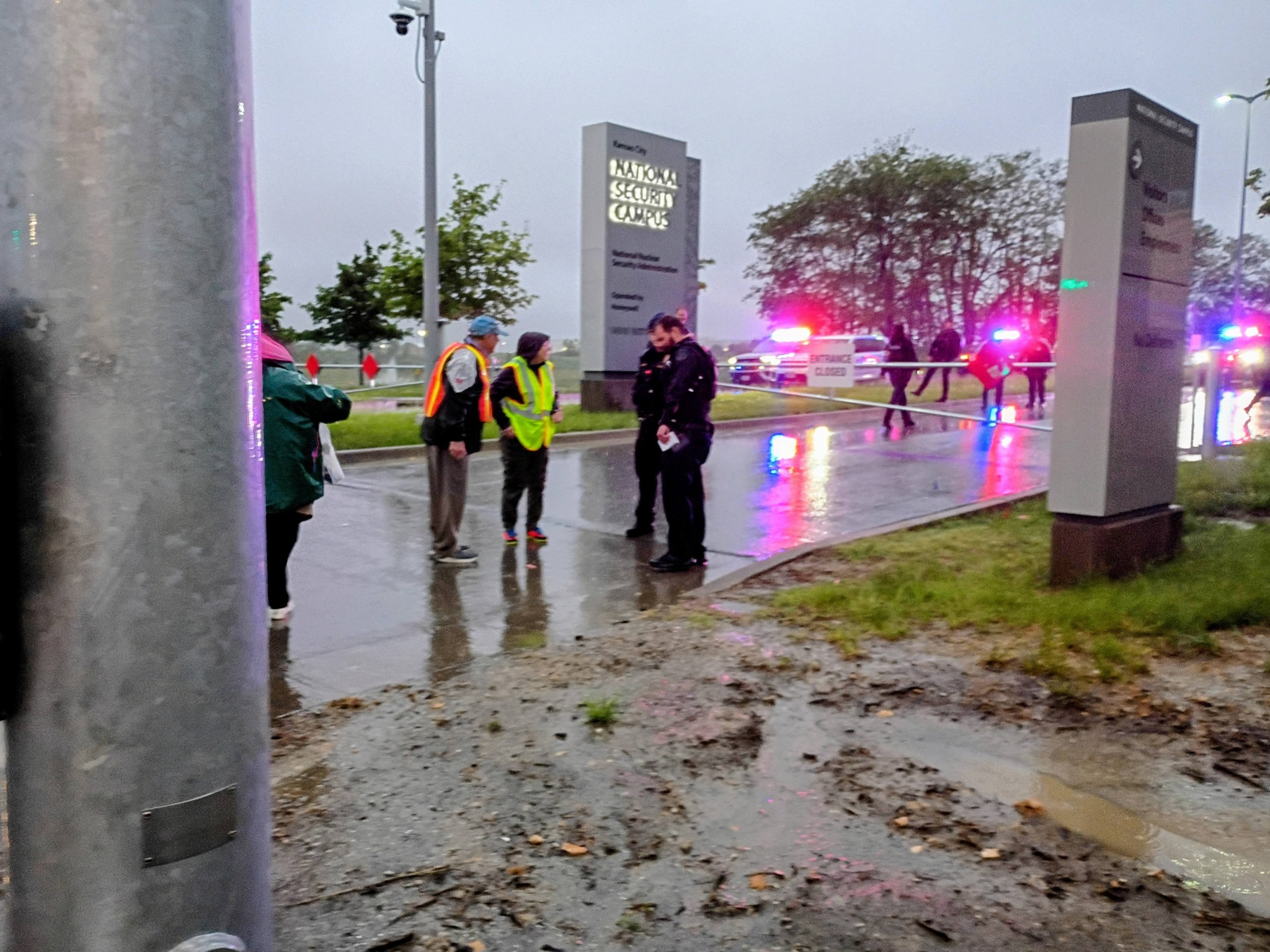 Police and vested activists stand on a wet roadway at dusk beside the lit "National Nuclear Security Campus" entrance sign, emergency lights flashing.