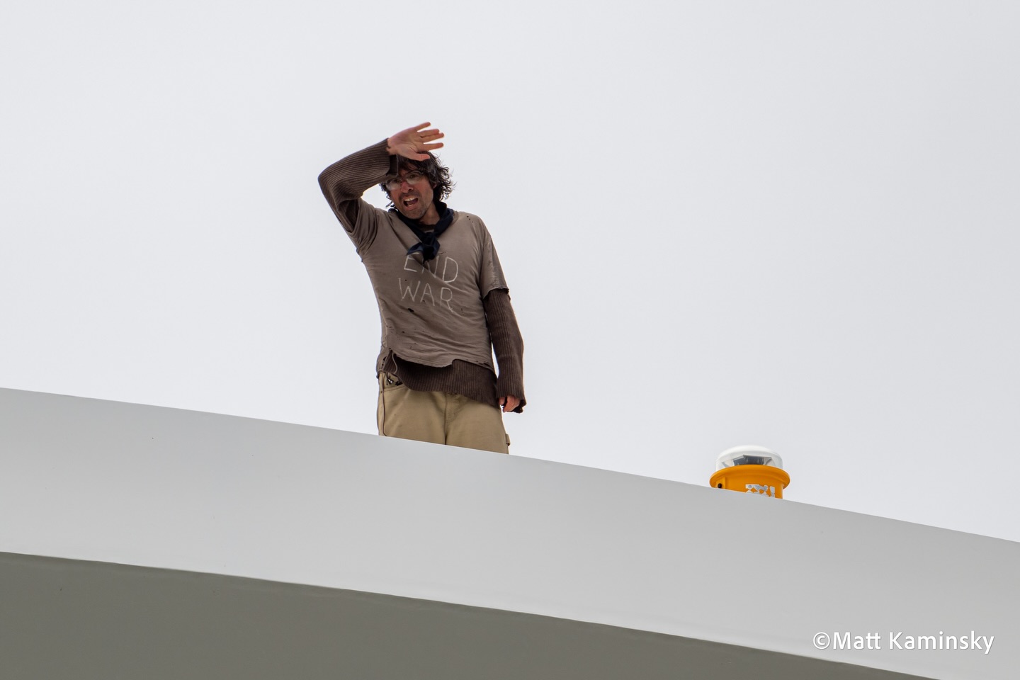 A man in a tan shirt stands atop the white curved span of a bridge, shielding his eyes with one hand against a grey sky.