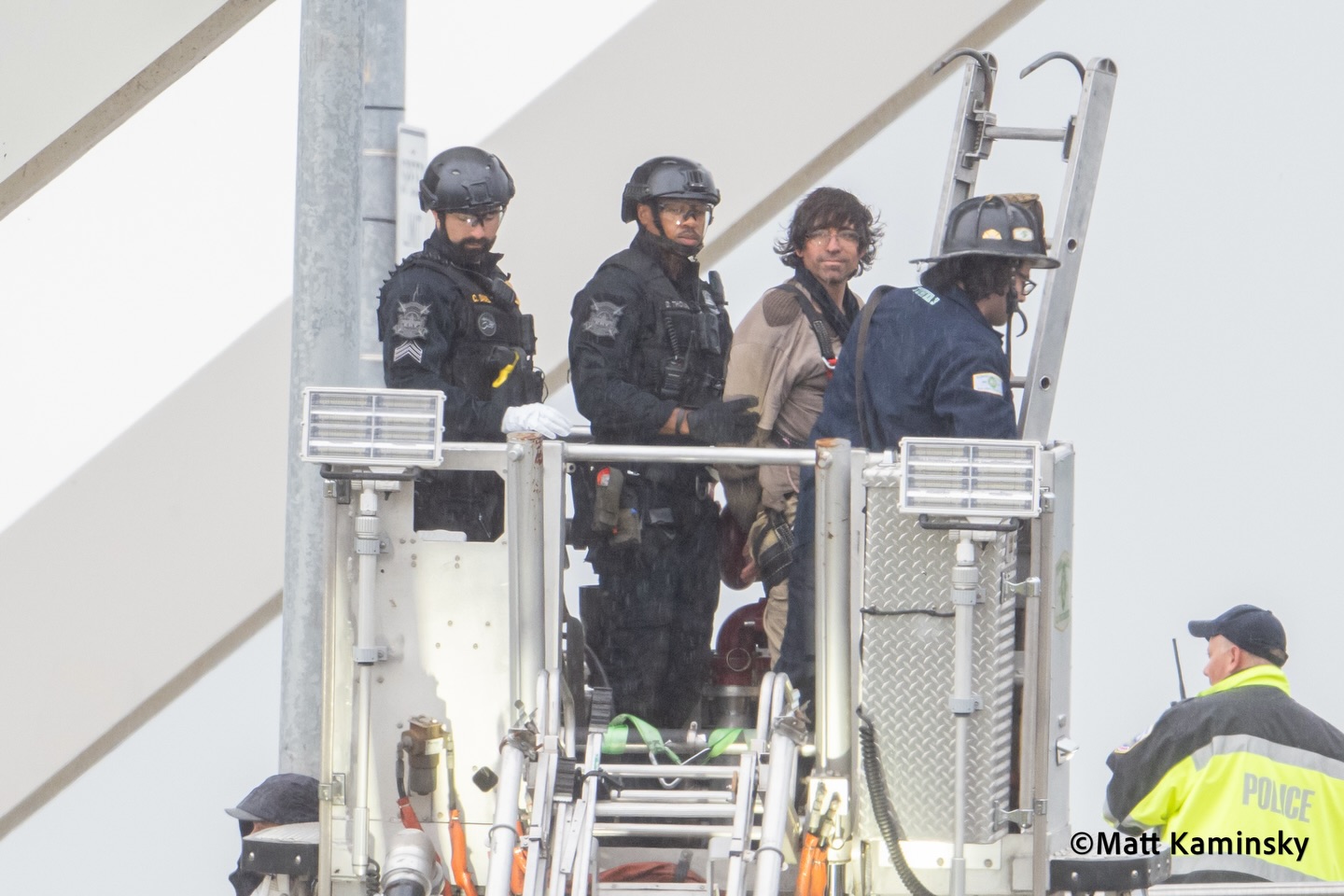 Helmeted tactical officers on a raised fire-truck bucket platform detain a man in a green jacket high above a bridge.