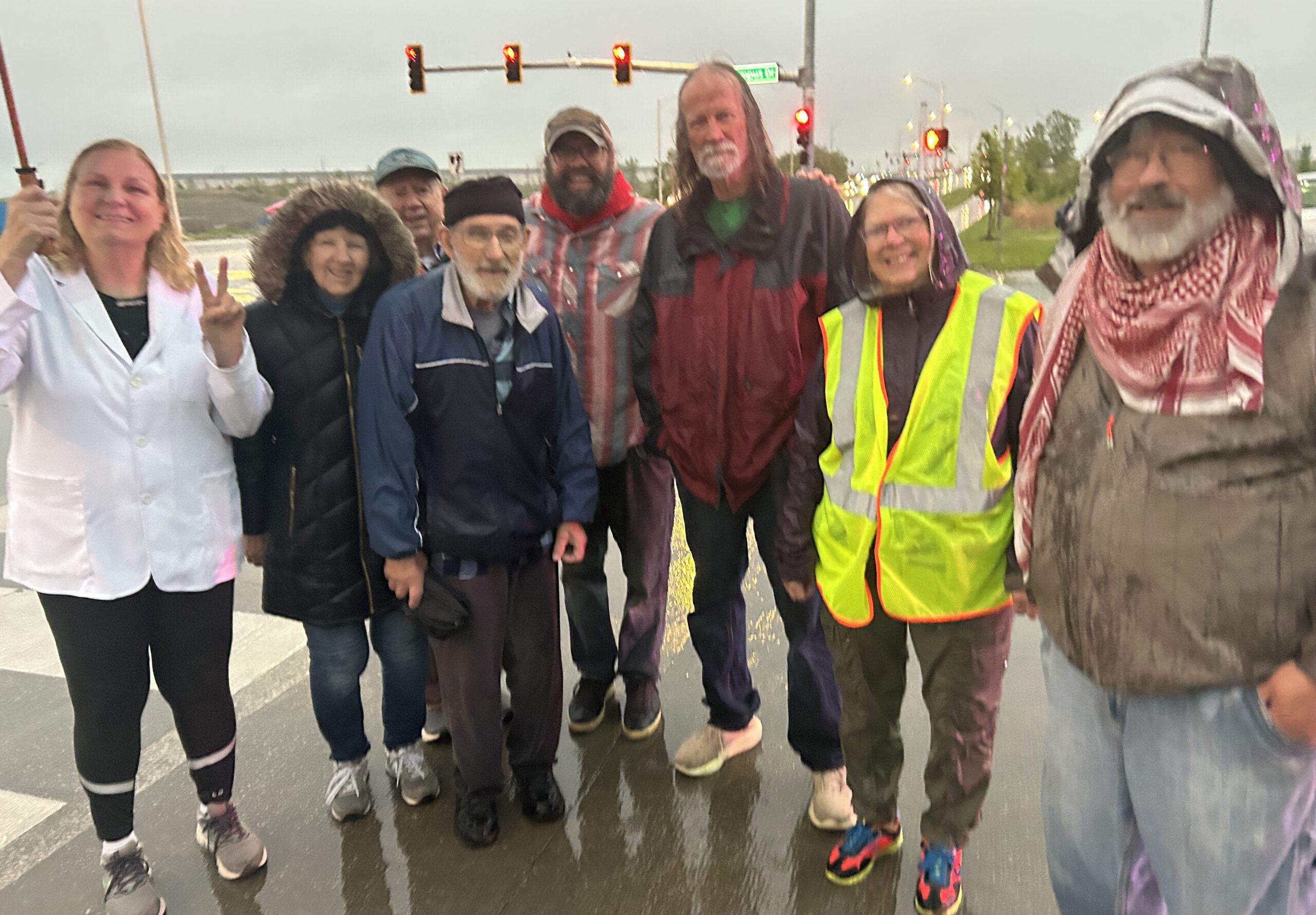 Seven activists in rain gear and safety vests stand together smiling at a roadside crossing under a traffic light.