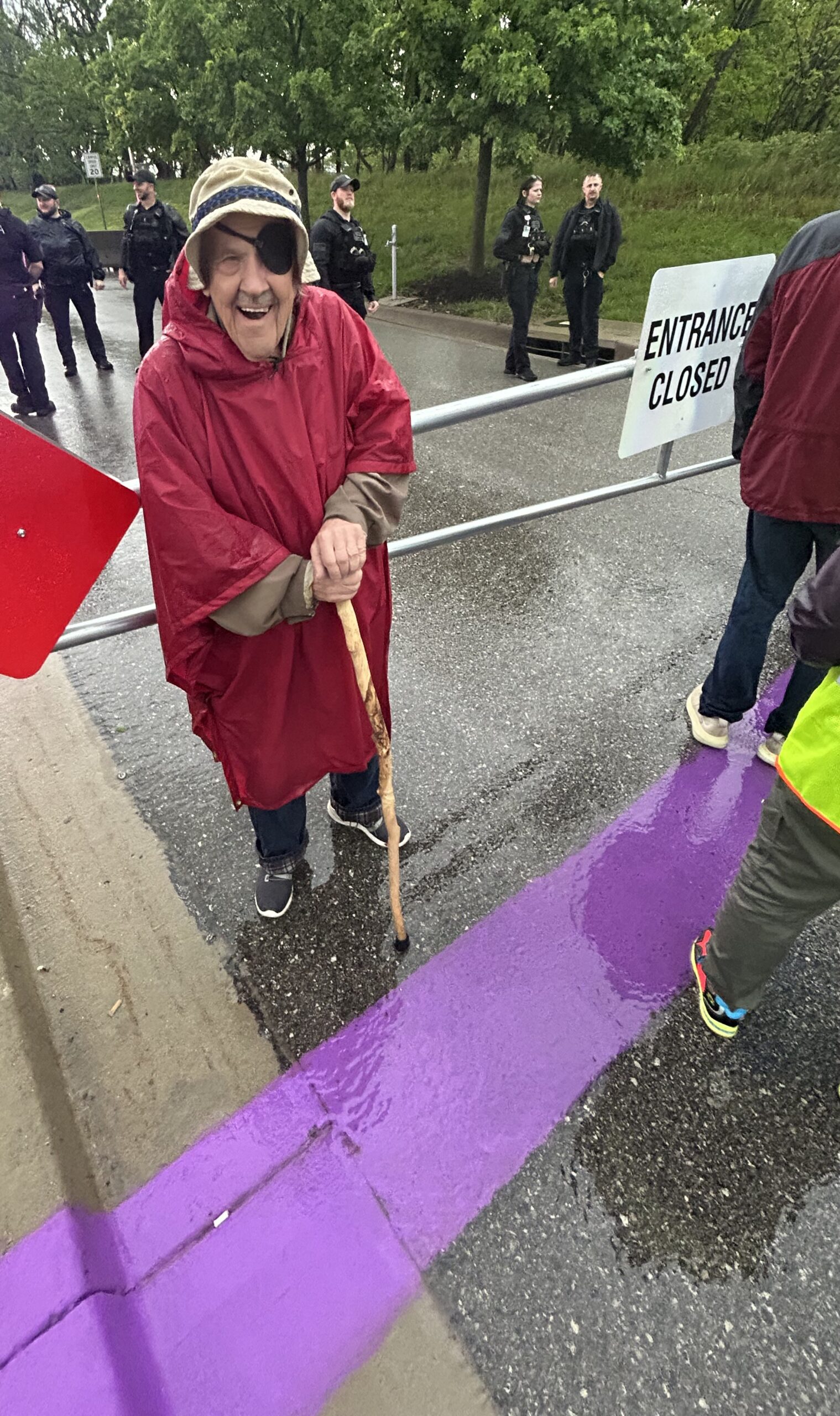 An older activist in a red rain poncho smiles while standing over a wide purple-painted stripe poured across wet pavement, holding a stick; an "ENTRANCE CLOSED" sign is behind.