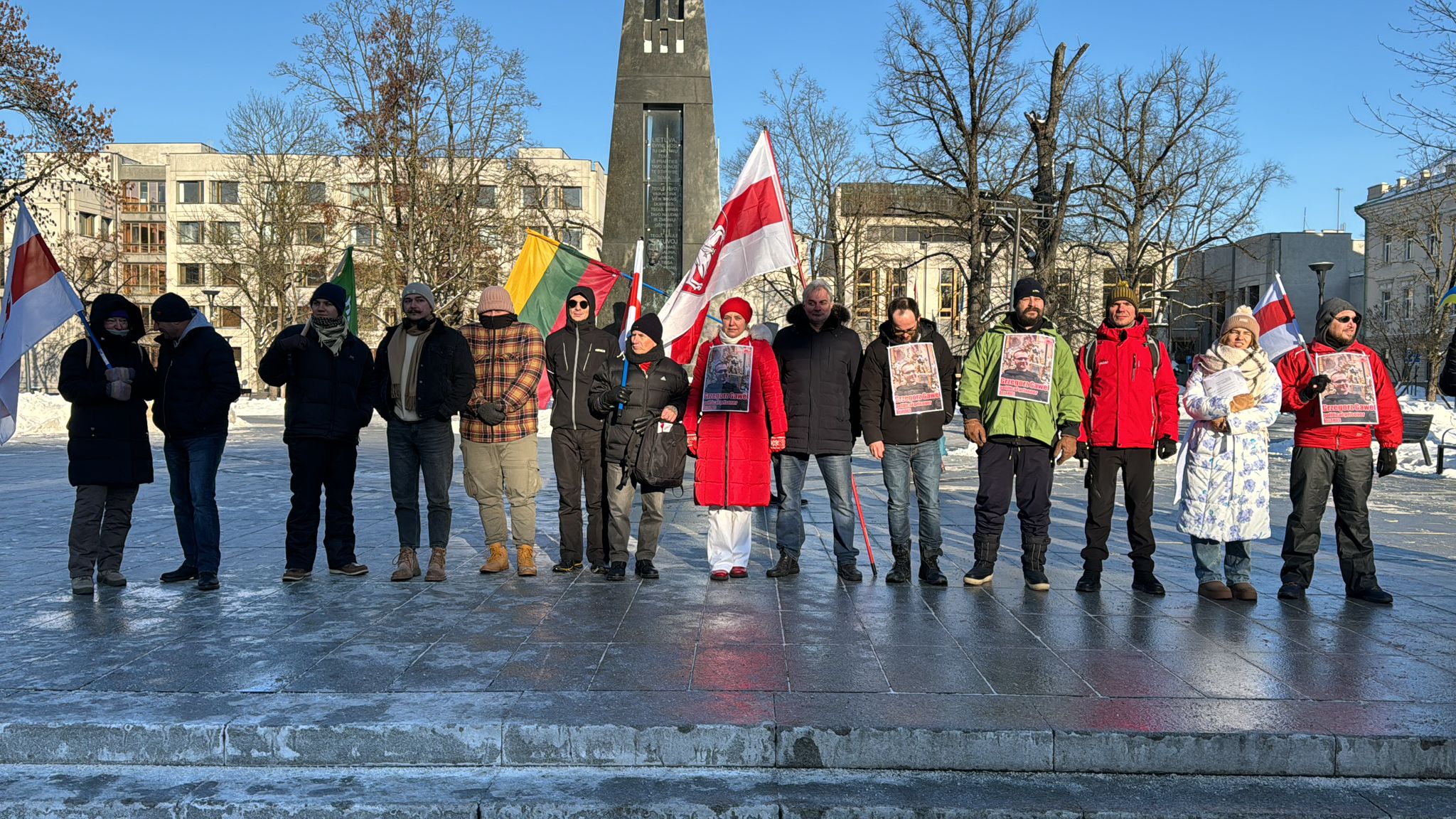 A line of people stand on an icy plaza in winter holding white-red-white Belarusian opposition flags and protest posters, a church tower behind.