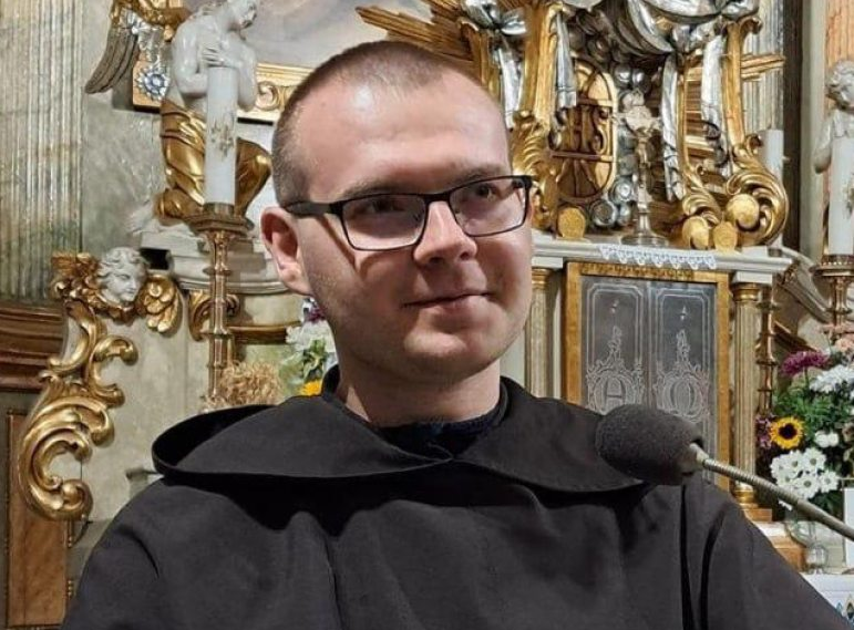 Portrait of a young Franciscan friar in a brown habit, wearing glasses, seated in front of an ornate gilded church altar.