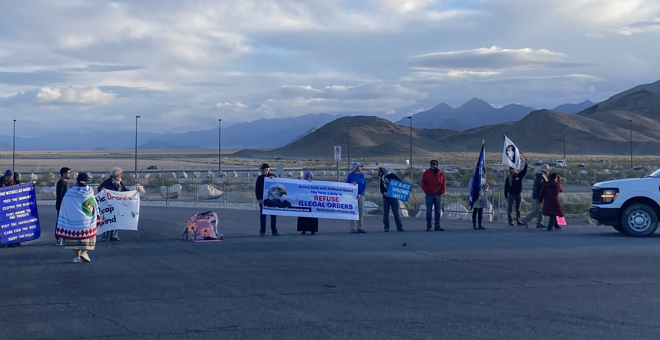 Activists stand across a desert parking area at dusk holding banners including "REFUSE ILLEGAL ORDERS" and "REAP THE WIND," mountains behind; a white truck with flashing lights is at right.