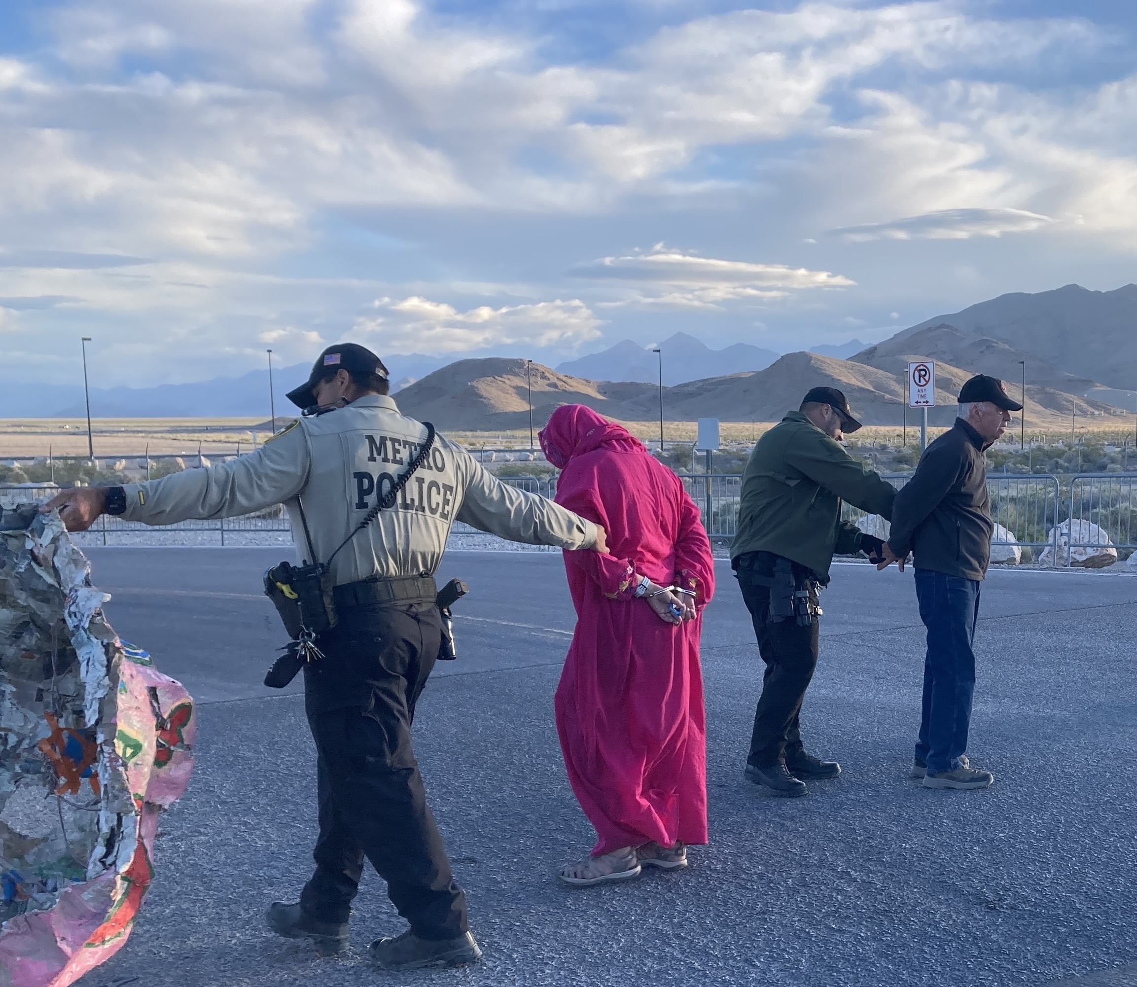 At dawn in the desert, police officers handcuff and lead away a person dressed in a long pink robe alongside other detained activists.
