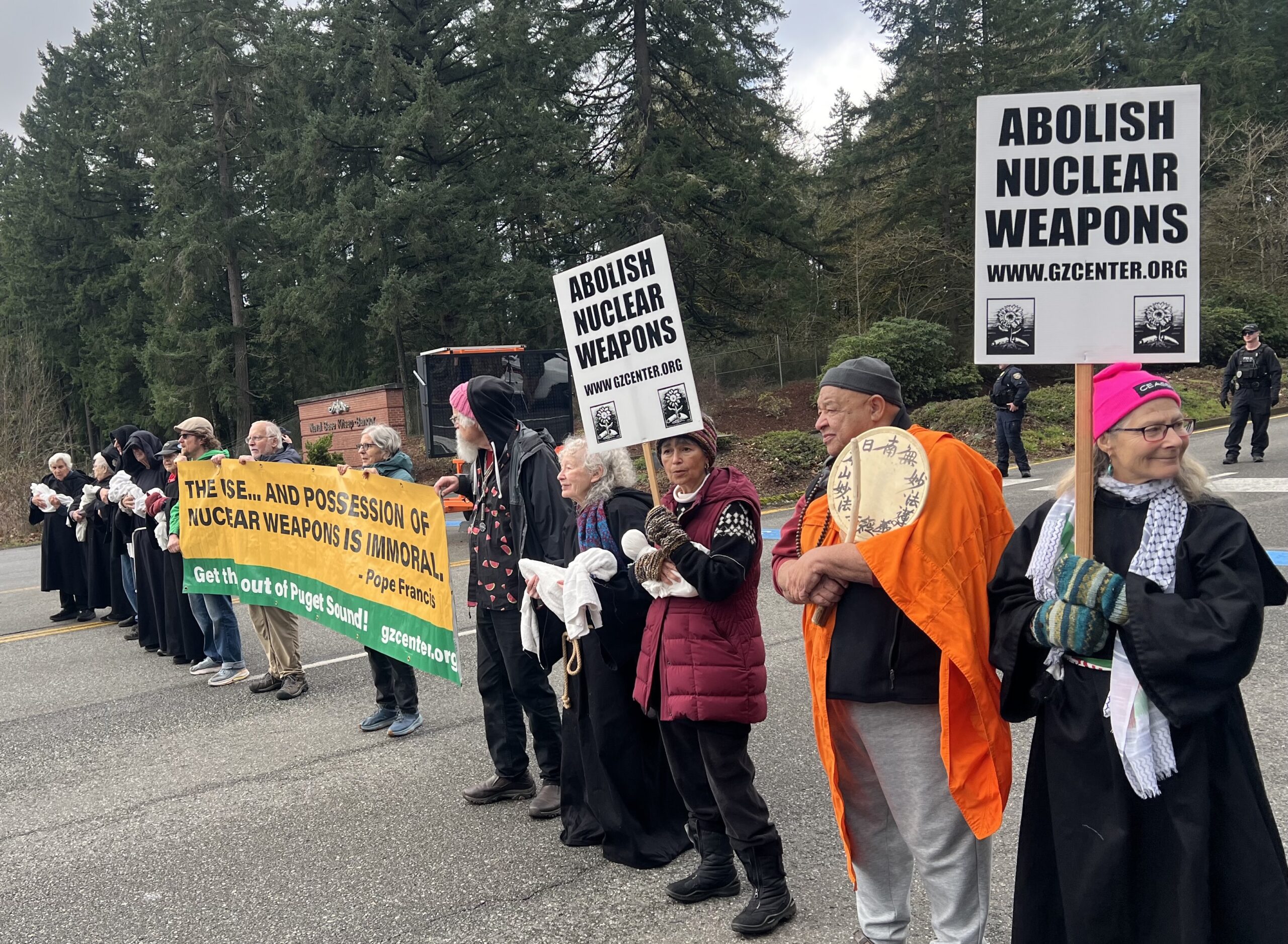 A line of activists, including a Buddhist monk in orange robes, hold "ABOLISH NUCLEAR WEAPONS" signs and banners along a roadside.