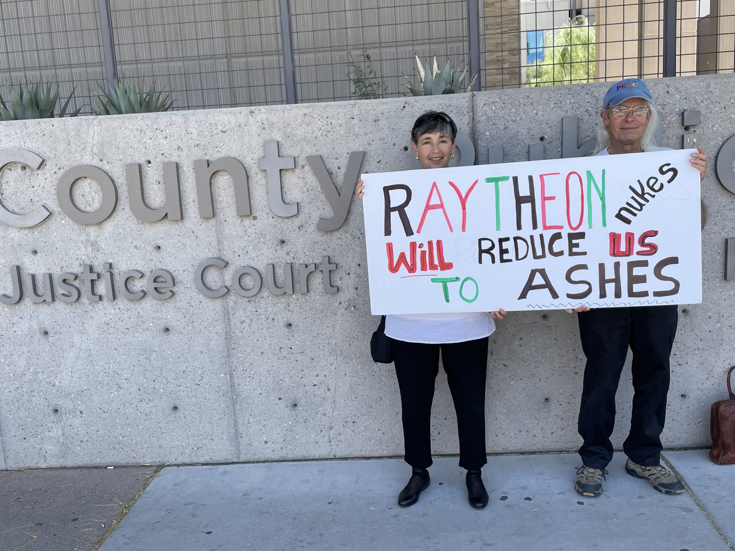 Two activists stand in front of a "County Justice Court" building holding a hand-lettered sign reading "RAYTHEON nukes WILL REDUCE US TO ASHES."