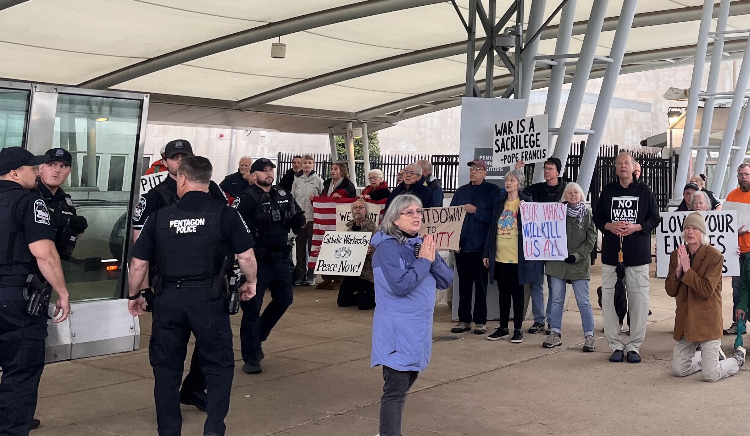 A crowd of protesters holds signs ("WAR IS A SACRILEGE – POPE FRANCIS," "YOUR WARS WILL KILL US ALL," "NO WAR," "LOVE YOUR ENEMIES," "Catholic Worker — Peace Now!") at a covered Pentagon entrance as Pentagon Police stand in front.