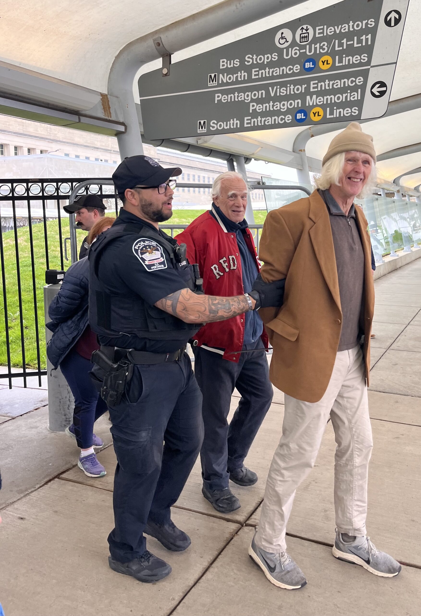 An officer in a "POLICE" vest handcuffs an older man as a younger man in a tan coat is also led away at a Metro/Pentagon entrance with directional signage behind.