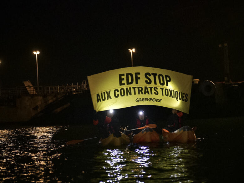 At night on the water, Greenpeace activists in small boats hold a lit banner reading "EDF STOP AUX CONTRATS TOXIQUES" in front of a docked ship.