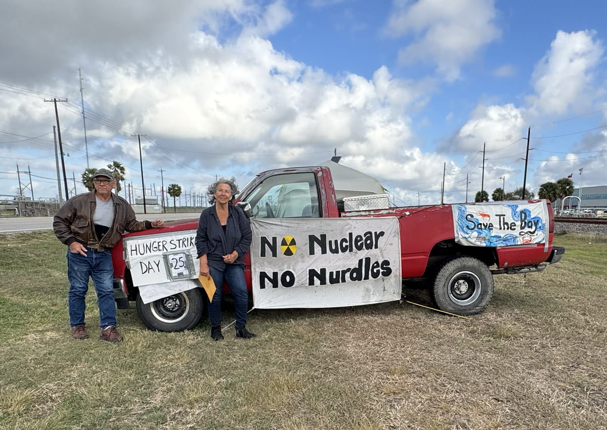 Two activists stand beside an old red pickup truck in a field with signs reading "NO Nuclear / No Nurdles" and "Save the Bay."