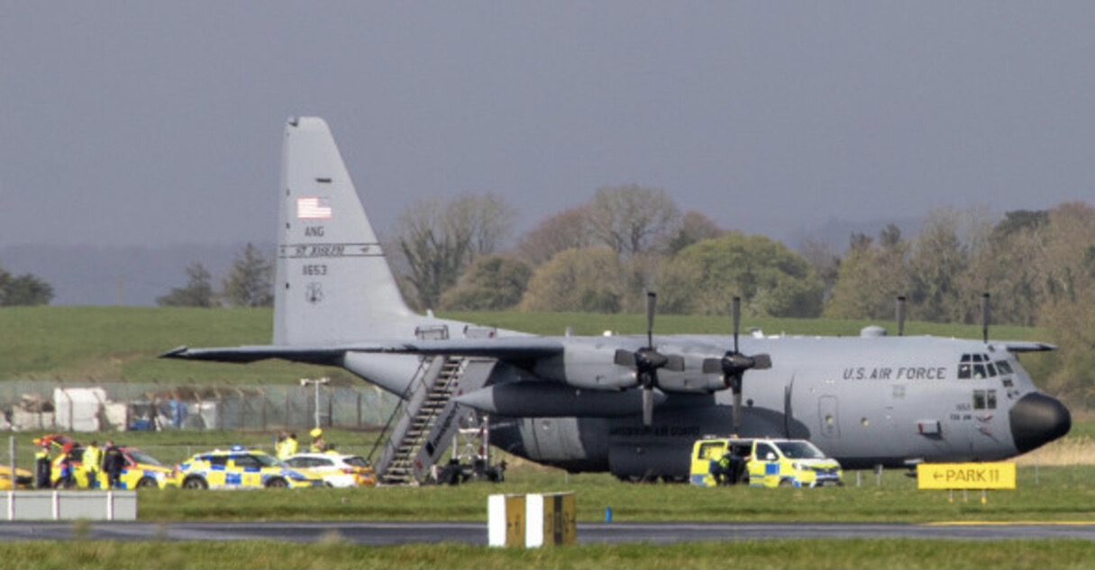 A grey U.S. Air Force C-130 transport plane parked on an airfield with boarding stairs deployed; police cars and emergency vehicles gathered beside it near a "PARK 11" sign.