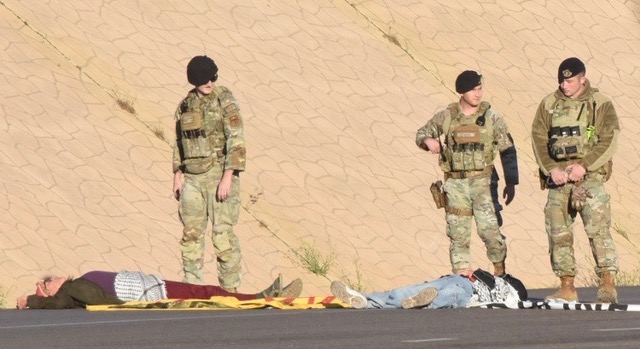 Two activists lie on the road in front of a desert base wall while three camouflaged military police in berets stand over them.