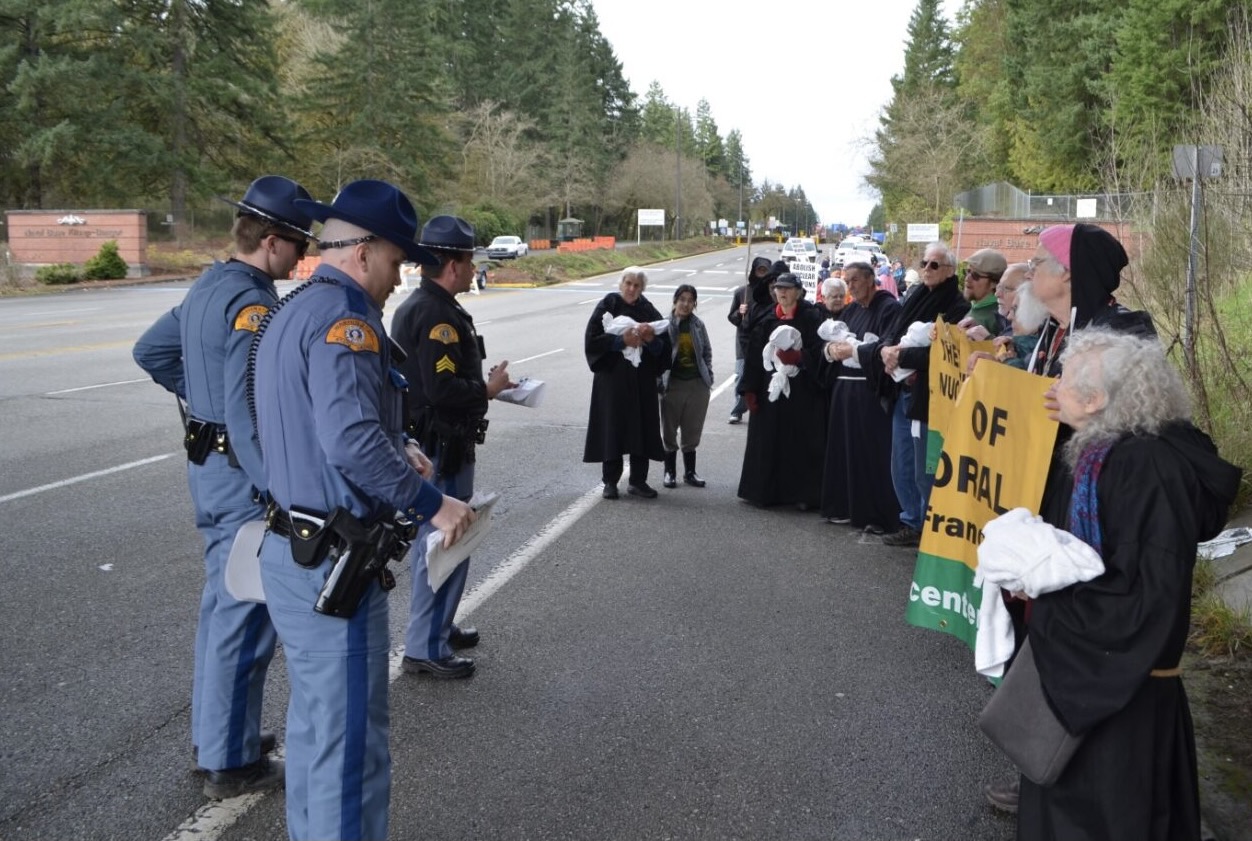 State troopers in campaign hats stand in a line across a road facing robed and bundled-up activists holding banners at a base entrance.
