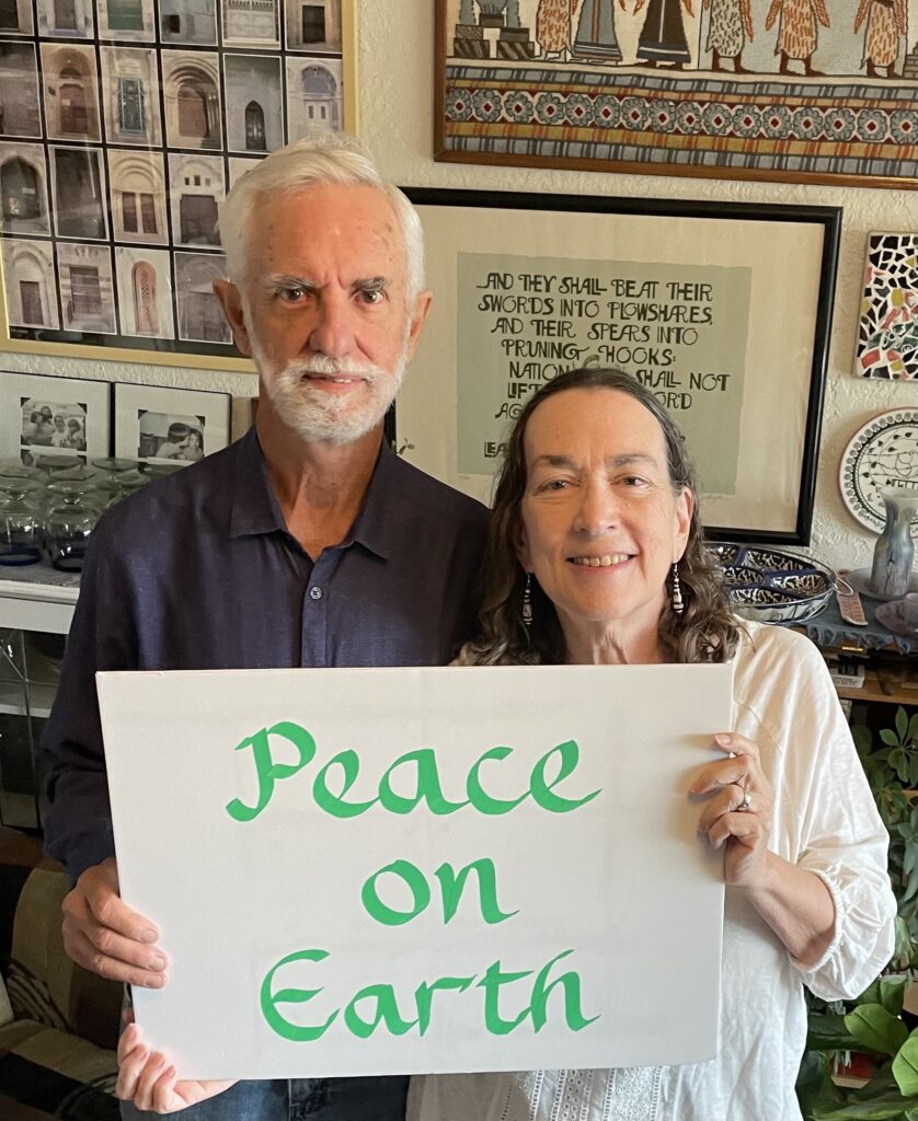 Jack and Felice Cohen-Joppa standing together at home, holding a hand-lettered sign that reads “Peace on Earth.” Framed on the wall behind them is a calligraphy of the passage from Isaiah about beating swords into plowshares.